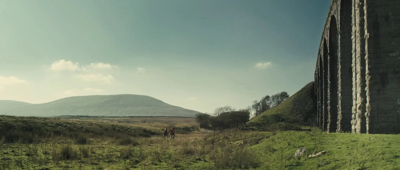 Sightseers (2012) movie still: wide — A vast, rolling green landscape features a large, stone arched viaduct on the right. Two people walk across…