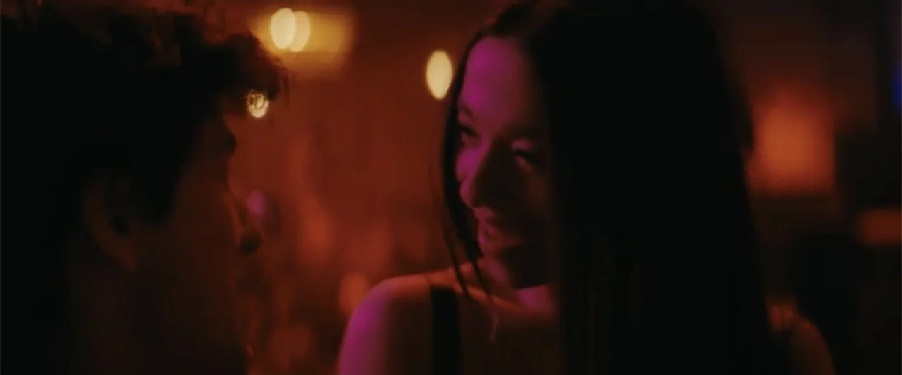 Anora (2024) movie still: closeup — A dimly lit club interior with soft out-of-focus background lights. A young woman turns her head to look at a…