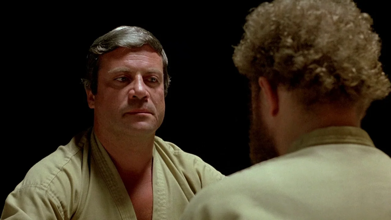 The Brood (1979) movie still: medium — A man in a martial arts gi sits in a dark, black-walled space. He faces another man whose back is turned to…