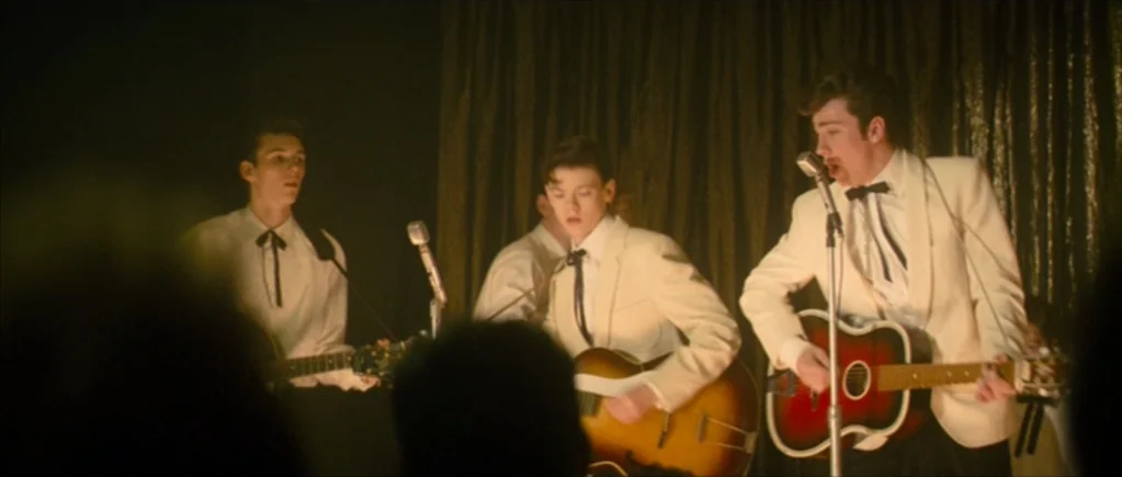 Nowhere Boy movie still: medium — A dimly lit indoor music stage features three men performing with guitars. Dark heavy curtains hang in the…