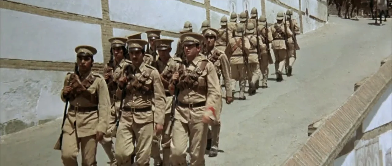 Duck, You Sucker (1971) movie still: fullbody — A group of colonial-era soldiers marches up a sloped dirt road beside a long wall. The men wear tan…