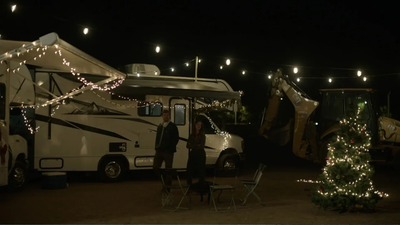 A Carpenter Christmas Romance (2024) movie still: wide — A dark outdoor space at night features an RV and construction machinery. A man and woman…