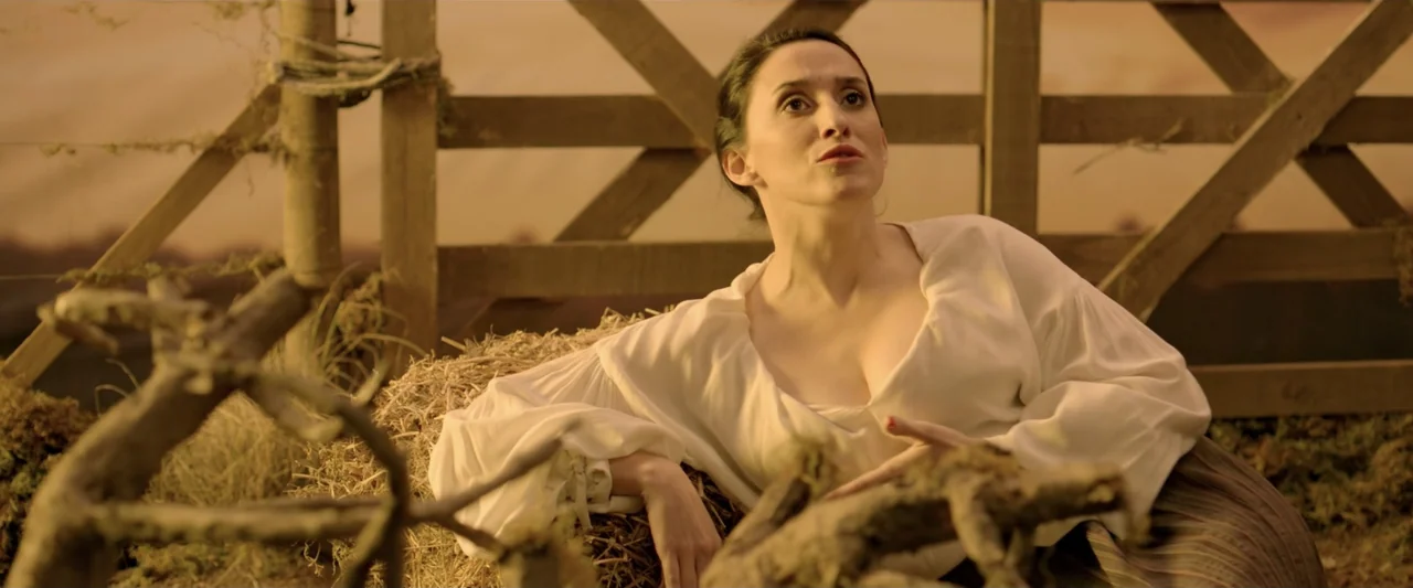 Perdida (2018) movie still: medium — A rural setting features a wooden fence behind stacks of dry hay bales. A woman in a white blouse reclines…