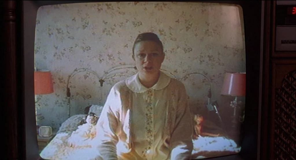 Mysterious Skin (2004) movie still: medium — A woman sits on a bed against floral wallpaper within a television screen frame. The subject faces the…