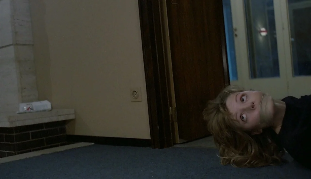 Angst (1983) movie still: closeup — A dark carpeted floor transitions into a doorway and a light-colored wall. A young woman lies on the ground with…