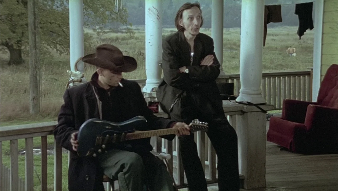 Hard Core Logo movie still: fullbody — A wooden porch with white pillars overlooks a grassy, open field during the day. Two men stand on the porch…