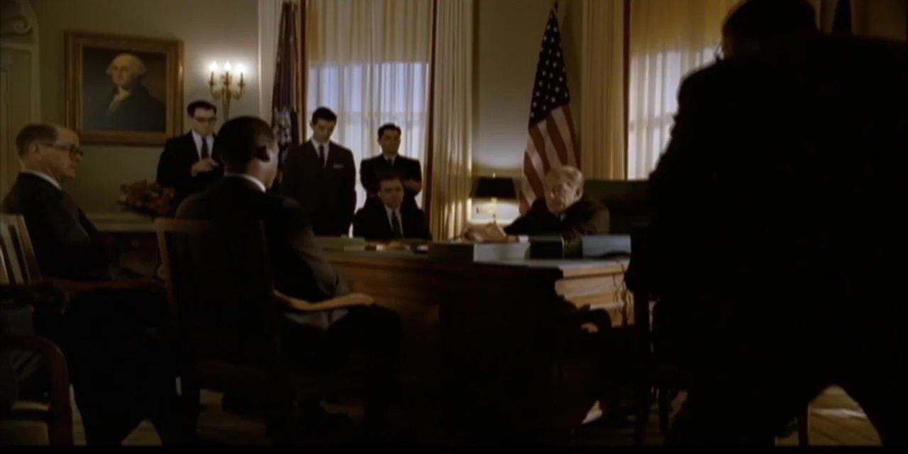 Path to War (2003) movie still: medium — A formal office with a large wooden desk and an American flag. A group of men in suits gather for a…