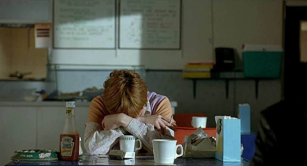 Butterfly Kiss (1995) movie still: medium — A quiet cafeteria setting features a menu board on the wall and table clutter. A woman sits alone at a…
