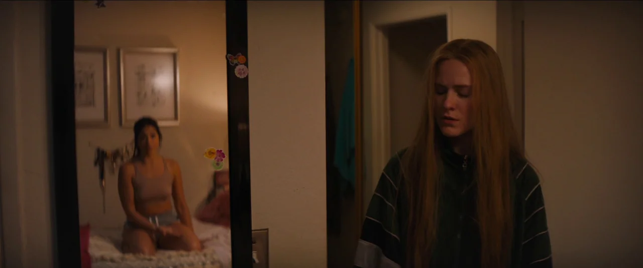 Kajillionaire (2020) movie still: medium — A dimly lit room contains a mirror showing a person on a bed. One woman faces forward in the dark…