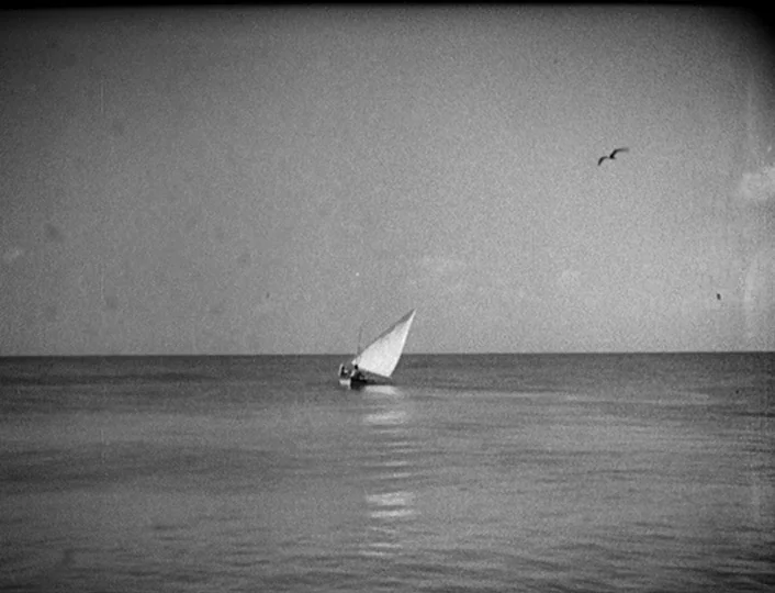 The White Sheik (1952) movie still: wide — A calm, vast ocean extends to a flat horizon line under an overcast sky. A small single-masted sailboat…