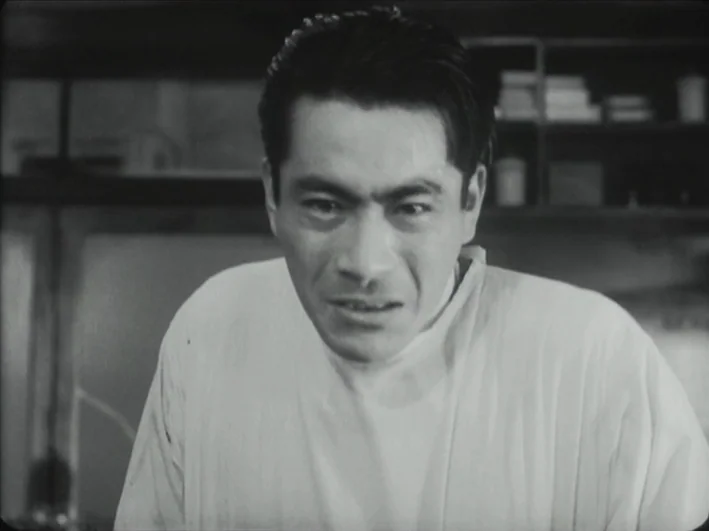 The Quiet Duel (1949) movie still: medium — A dimly lit room with medical shelves visible in the blurred background. A man in a white surgical gown…