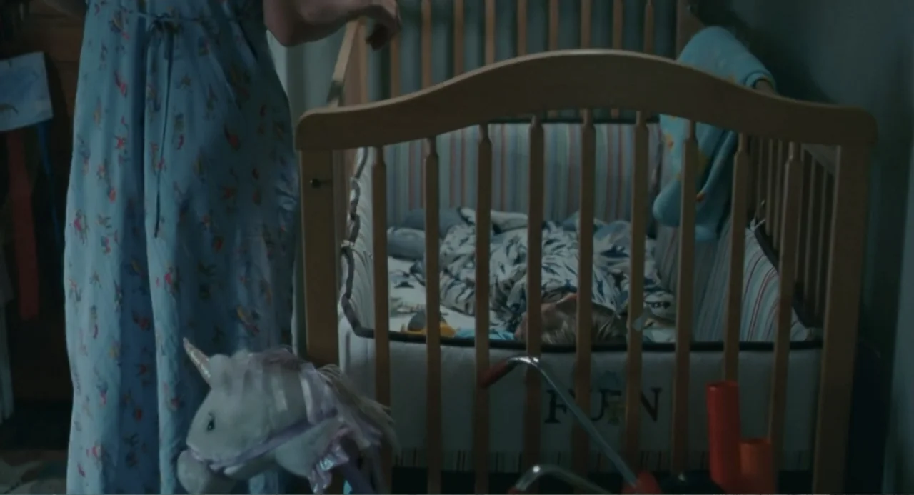 Motherhood (2020) movie still: medium — A dark, quiet nursery features a wooden crib and a plush unicorn toy nearby. A person stands beside the crib…