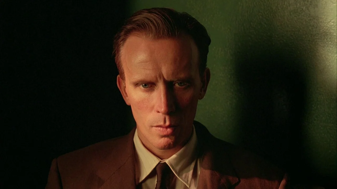 Naked Lunch (1991) movie still: closeup — A man in a brown suit occupies a dim room with green wall paint. He looks directly at the camera with a…