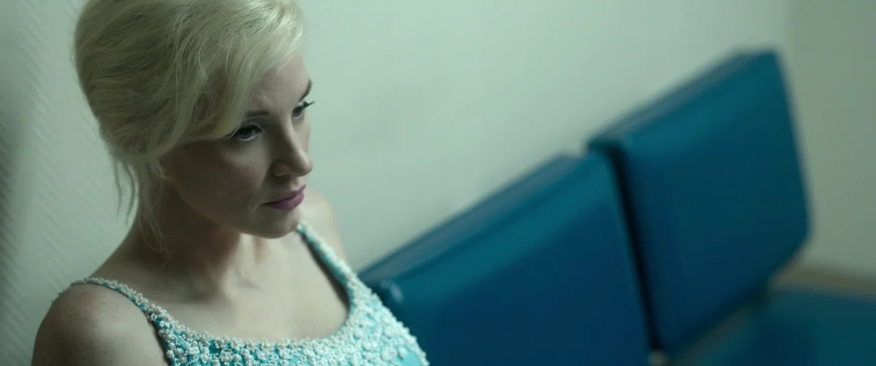 Mother’s Instinct (2024) movie still: medium — A woman sits on a blue vinyl bench against a pale green wall. She stares forward while wearing an…