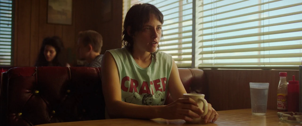 Love Lies Bleeding (2024) movie still: medium — A diner booth with red vinyl upholstery and wood paneling walls. A woman sits alone holding a coffee…