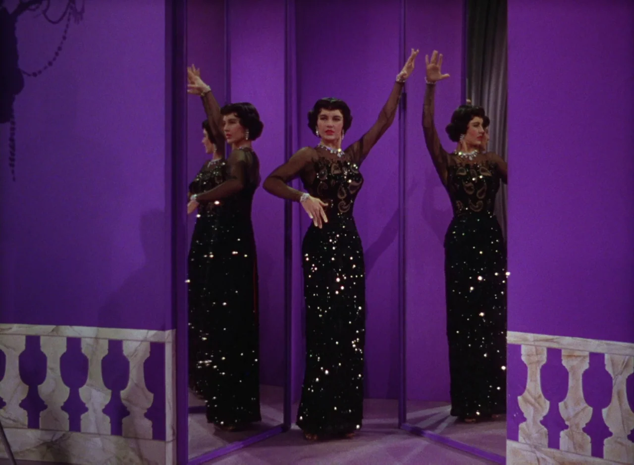 The Band Wagon (1953) movie still: fullbody — A woman stands in a bright purple room featuring large mirrors and white balustrades. She strikes a…