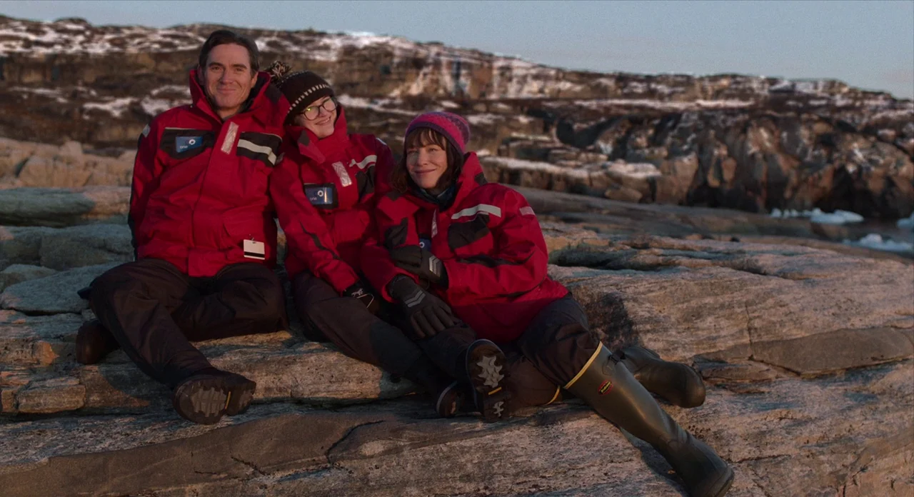 Where’d You Go Bernadette (2019) movie still: fullbody — Three people sit close together on a large, lichen-covered rocky surface near water. A…
