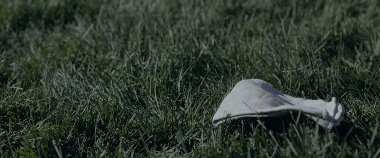 Radius (2017) movie still: closeup — A patch of dark green grass fills the frame outdoors under natural daylight. A crumpled white cloth rests…