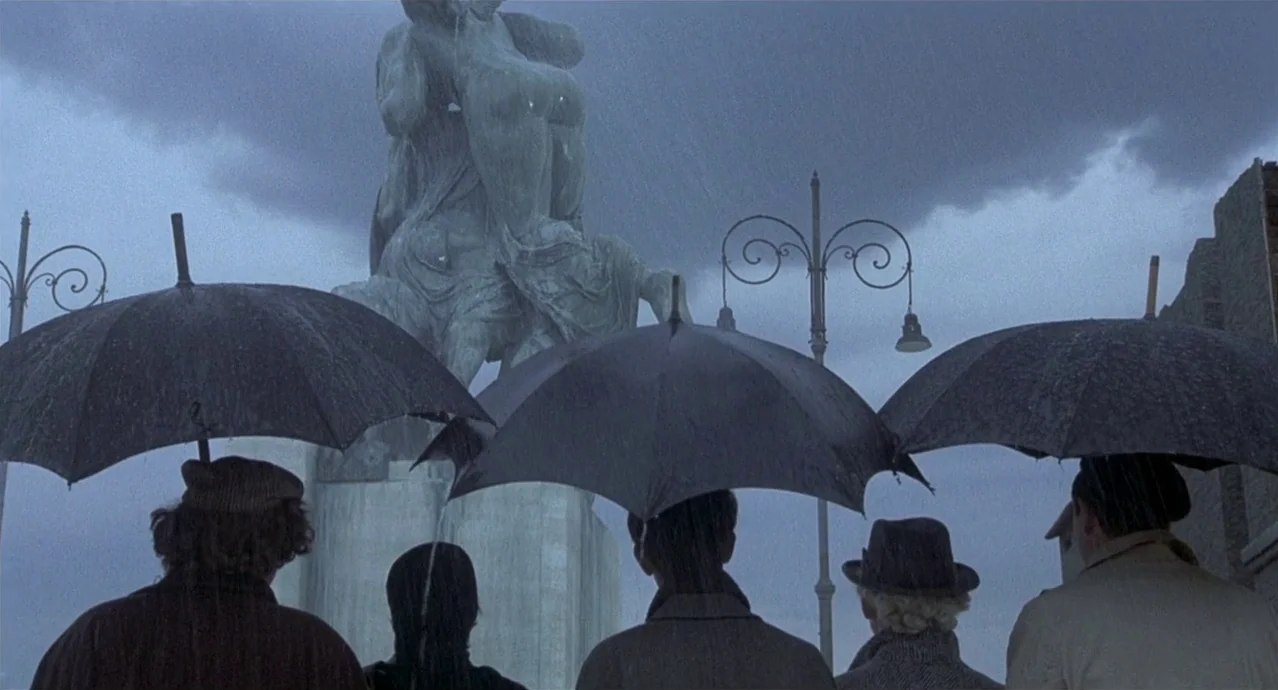 Amarcord (1973) movie still: wide — A group of people standing under umbrellas in a rain-swept city square. They gaze up toward a large stone…