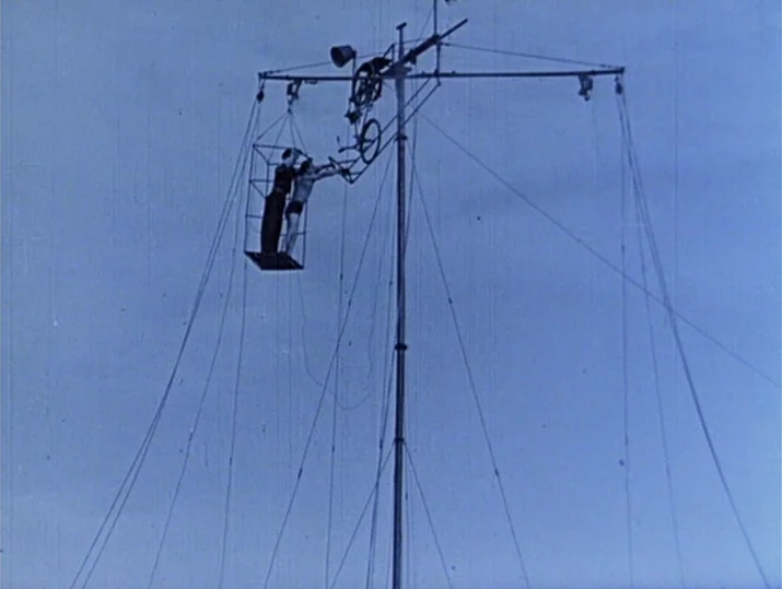 Innocence Unprotected (1968) movie still: wide — A high, complex circus apparatus stands against a clear, expansive blue sky. Two acrobats perform a…