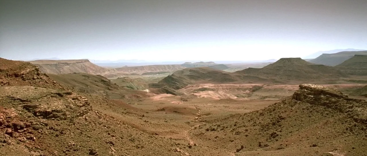 The Hills Have Eyes (2006) movie still: establishing — An expansive arid desert landscape features rolling mesas and dry, rocky terrain. Sparse…