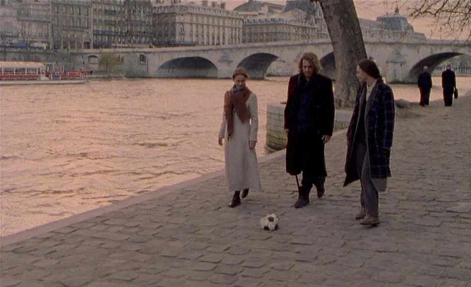 Pola X (1999) movie still: fullbody — A stone embankment beside a river features a cobblestone surface and a large tree. Three individuals walk…