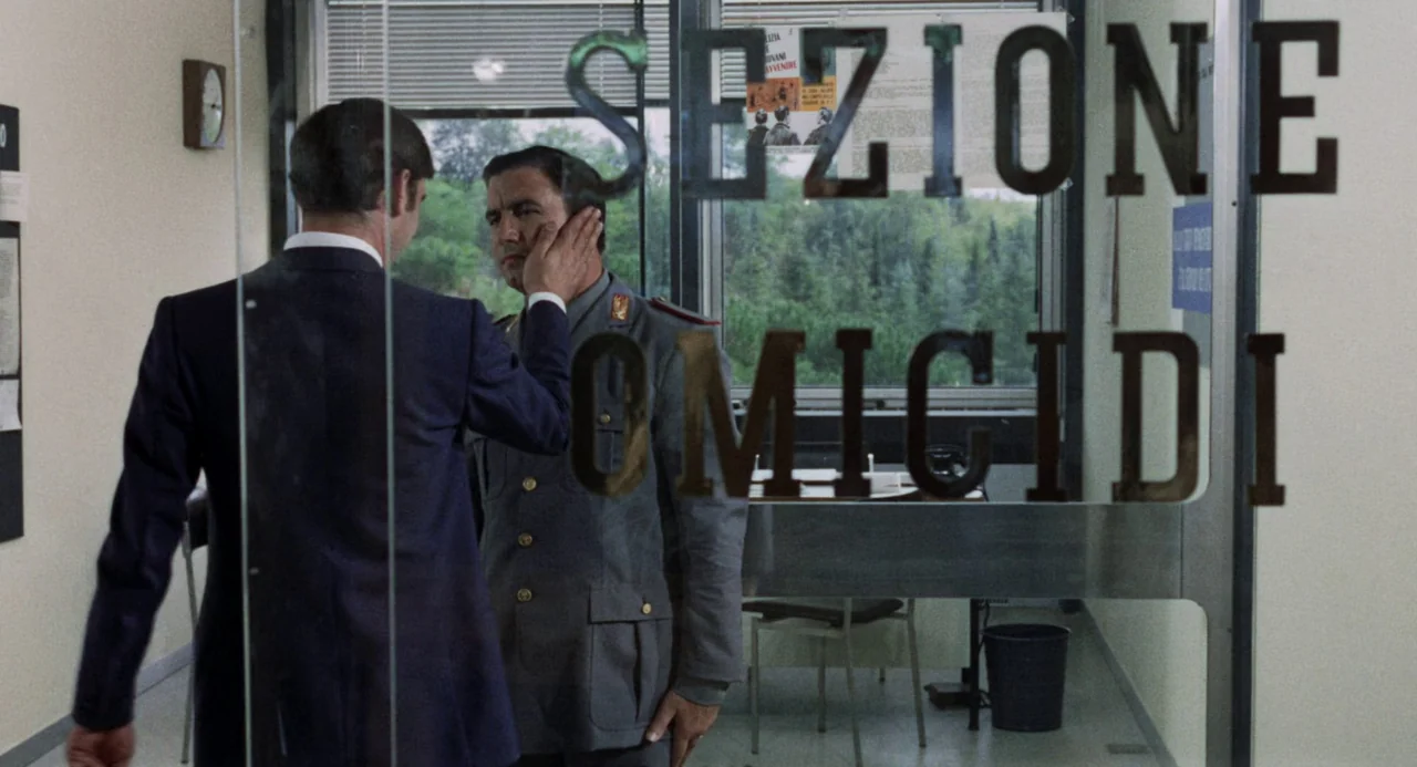 Investigation of a Citizen Above Suspicion (1970) movie still: medium — A police station office features a glass door with prominent black printed…