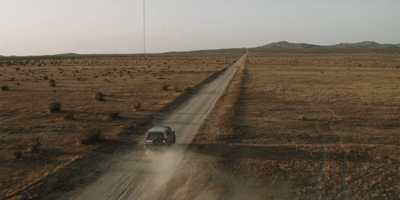 Fall (2022) movie still: wide — An arid, flat desert expanse is bisected by a long, straight dirt road. A lone vehicle drives away into the…