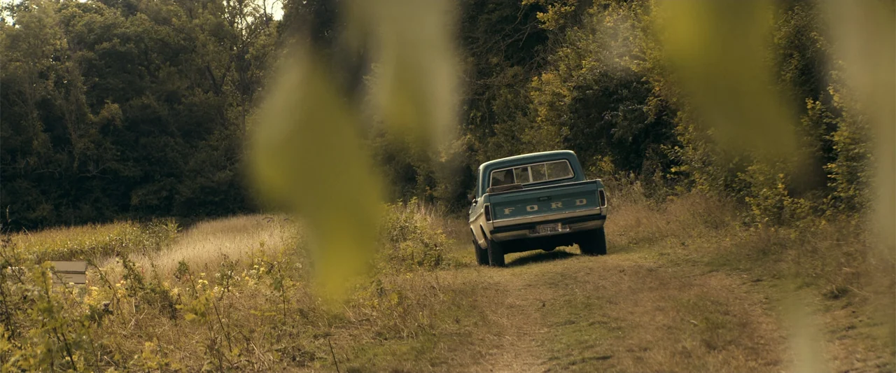 The Beekeeper (2024) movie still: wide — A teal vintage Ford pickup truck sits parked on a dirt path in a field. Tall trees and wild grass surround…
