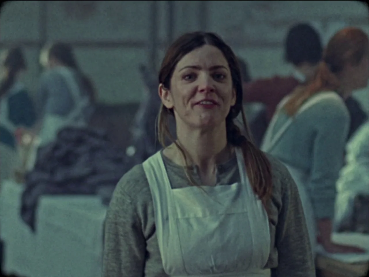 The Devil’s Doorway (2018) movie still: medium — A dimly lit, sparse room with muted gray walls and industrial-looking work tables. Several women in…