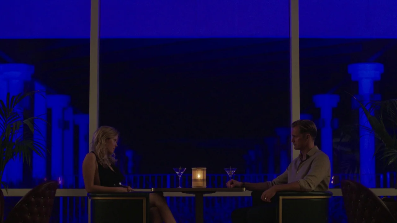 Infinity Pool (2023) movie still: wide — A modern interior space features a large window overlooking a brightly lit blue exterior. A man and a woman…
