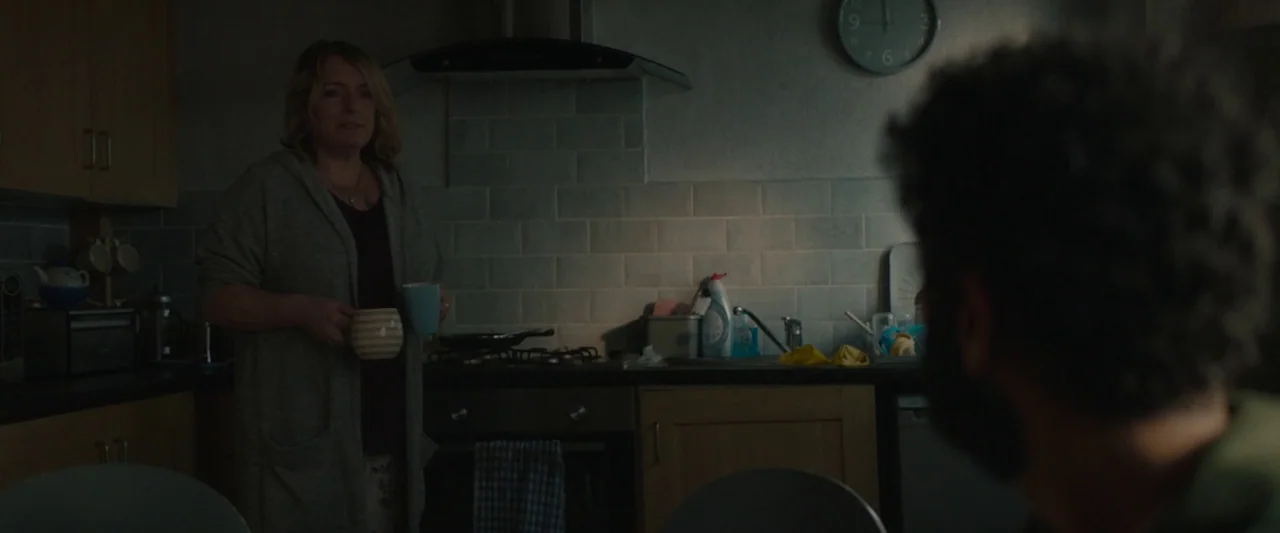 Ali & Ava (2021) movie still: medium — A dimly lit kitchen features wood cabinetry, a tiled backsplash, and a wall clock. A woman in a grey cardigan…