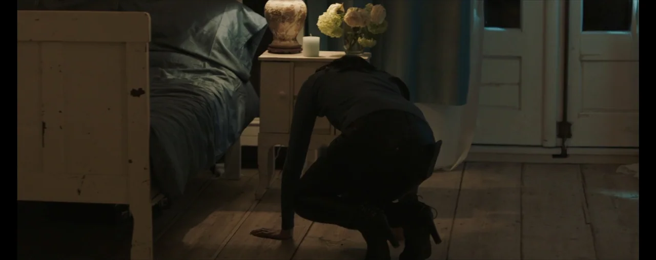 The Wrong Tenant (2022) movie still: medium — A dimly lit room with wooden floors contains a white bed frame and nightstand. A person in dark…