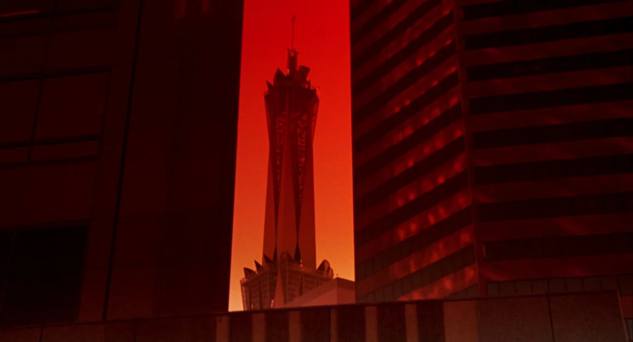 Night of the Comet movie still: establishing — A tall, slender tower stands between two dark skyscrapers against a bright red sky. The architecture…