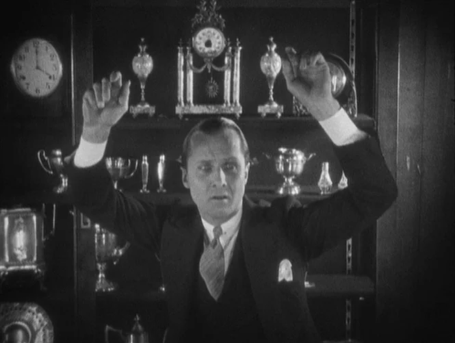 Underworld (1927) movie still: medium — A man stands before a shelf packed with various antique clocks and silver trophies. He holds his hands…