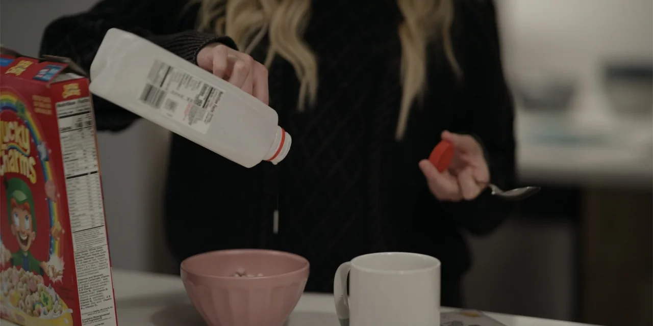 About Fate (2022) movie still: medium — A clean interior kitchen counter features a cereal bowl and a milk jug. A person in a black sweater pours…