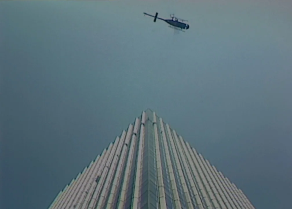 No (2012) movie still: establishing — A tall, tapered skyscraper reaches upward toward a cloudy, overcast sky. A small helicopter maintains a steady…