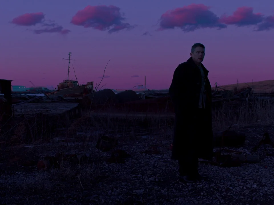 First Reformed (2017) movie still: fullbody — A desolate, industrial wasteland features the rusted hulls of abandoned ships near water. A lone man…