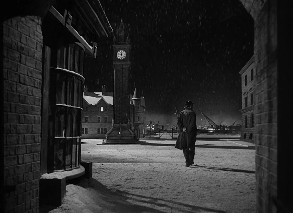 Odd Man Out (1947) movie still: wide — A snowy, deserted city square at night features a prominent central clock tower. A man in an overcoat walks…