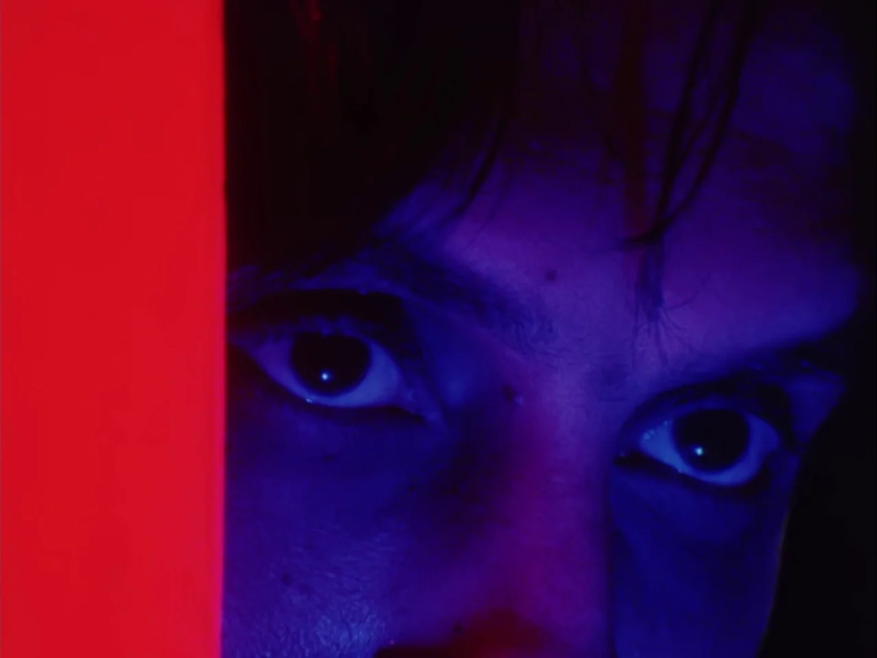 Subconscious Cruelty (2000) movie still: closeup — A close-up view shows a face illuminated by harsh red and blue light. The subject looks directly…