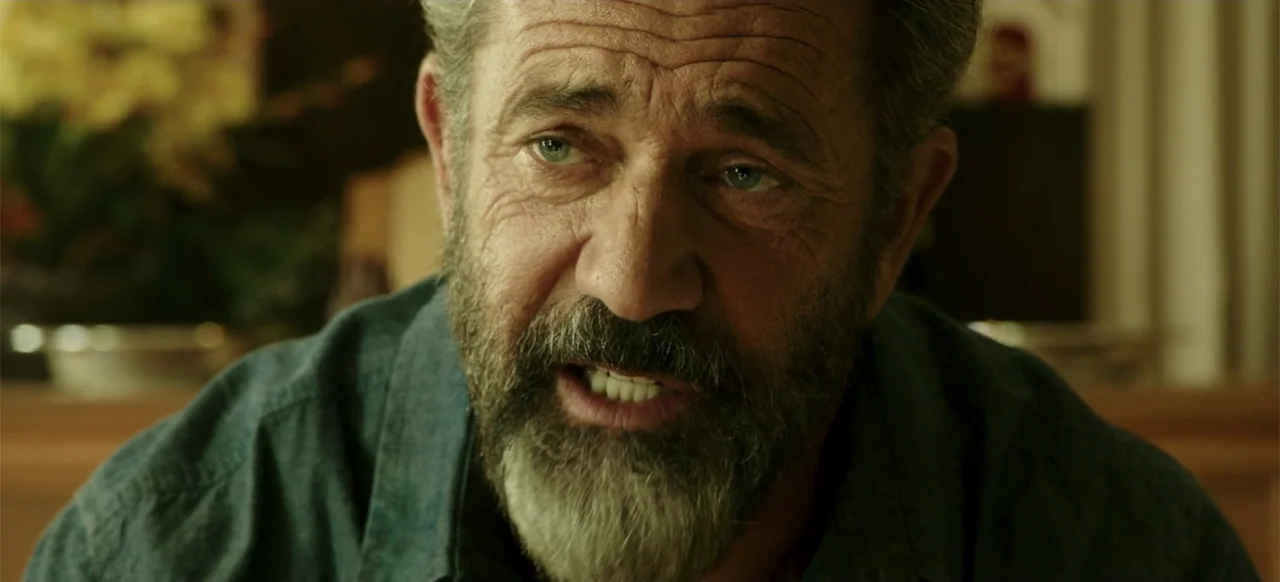 Blood Father (2016) movie still: closeup — An interior space with a dimly blurred background of neutral furniture and decor. A man with a gray beard…