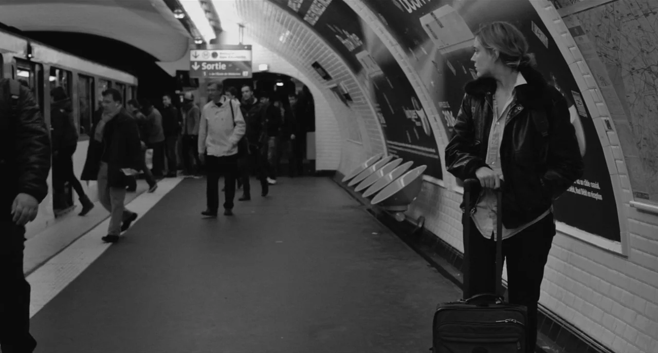 Frances Ha (2012) movie still: medium — A Paris metro station platform with curved tiled walls and large advertising posters. A woman stands beside…