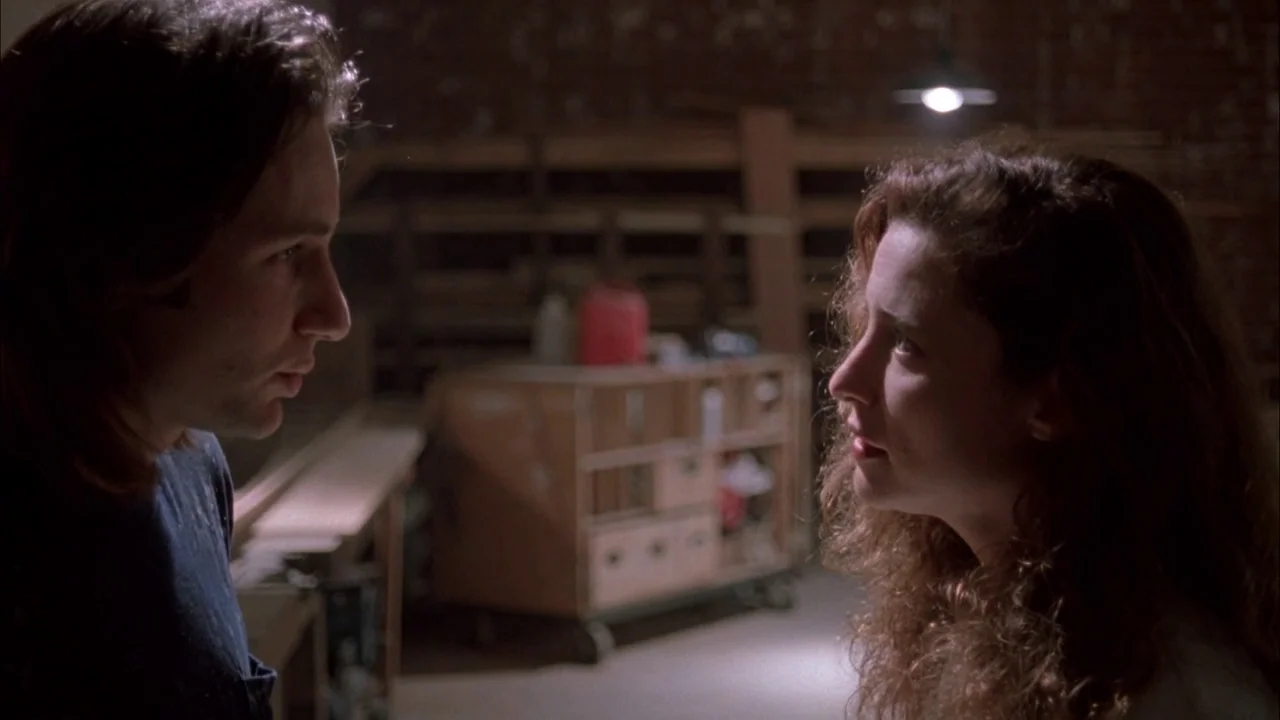 The Rapture (1991) movie still: medium — A dim workshop with a wooden cart and scattered tools in the background. A man and a woman stand facing…