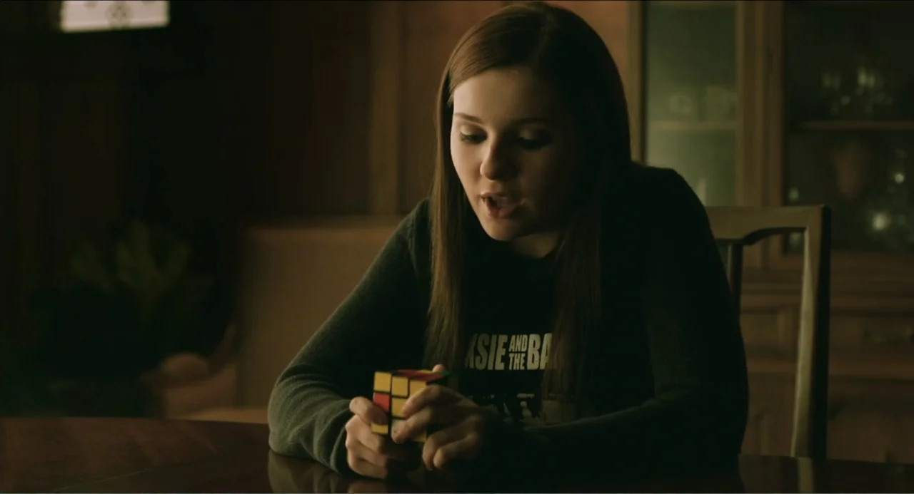 Haunter (2013) movie still: medium — A dimly lit room features a wooden dining table and a dark china cabinet. A young woman with long hair sits at…