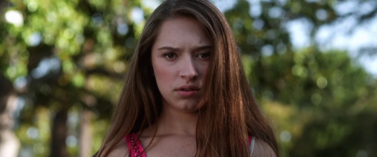 Excision (2012) movie still: closeup — A woman stands outdoors with out-of-focus trees and sunlight in the background. She faces the camera directly…