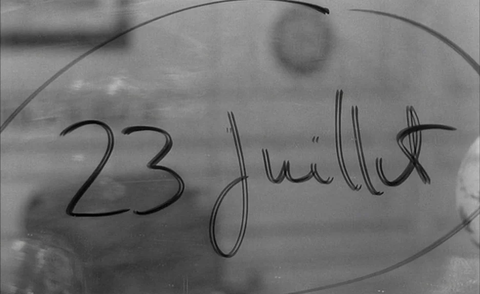 Le Feu Follet (1963) movie still: closeup — A glass pane features the handwritten date 23 juillet circled in black ink. The background shows blurred…