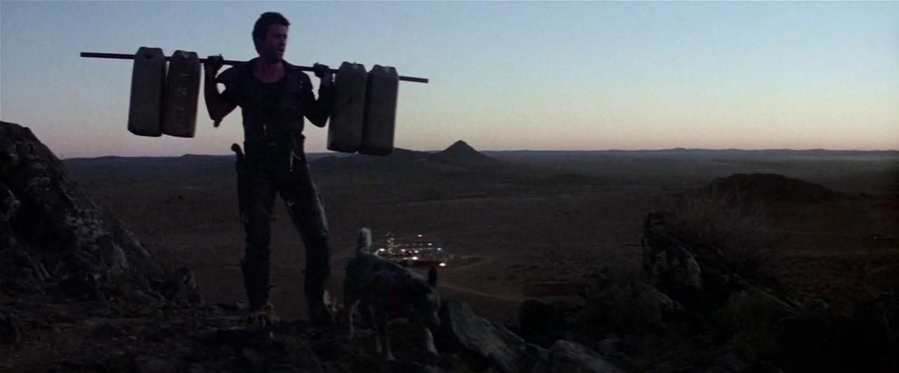 Mad Max 2 (1981) movie still: fullbody — An arid, rocky desert landscape stretches out under a dusk sky. A man stands on a ridge accompanied by a…