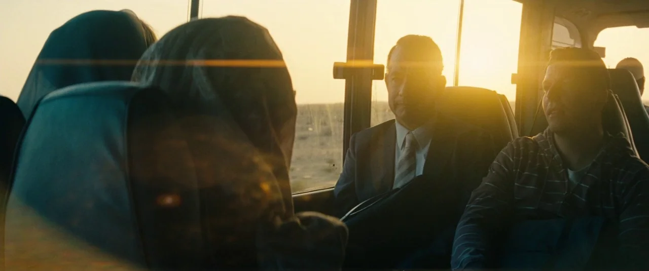 A Hologram for the King movie still: medium — Passengers sit inside a bus as it travels across a desert at sunset. The golden sun flares through the…