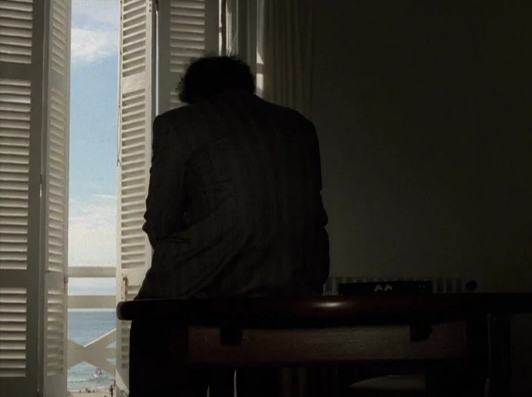 Keep Your Right Up (1987) movie still: medium — An interior space with a table sits in near darkness. A person sits silhouetted against an open…
