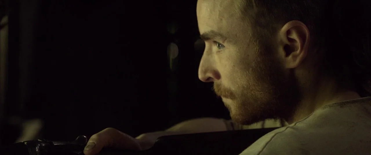 The Survivalist (2015) movie still: closeup — A dimly lit cabin of a vehicle at night with deep shadows surrounding the passenger. A man with a…